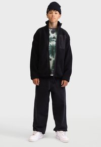 Black fleece jacket with zip, textured fabric, and a chest pocket; layered over a green tie-dye shirt and worn with black baggy jeans and white sneakers.