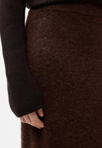 Maglione lavorato a maglia marrone con maniche lunghe, abbinato a una gonna di feltro marrone. Materiali testurizzati creano un effetto a strati.