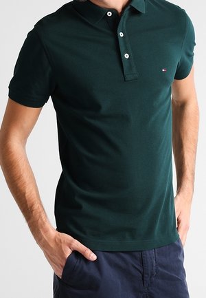 Mann trägt ein dunkelgrünes kurzärmeliges Polo-Shirt mit kleinem Logo auf der Brust und dunkelblaue Hose, Hände in den Taschen.