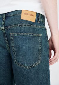 Vue arrière d'une personne portant un jean en denim bleu avec un patch en cuir beige portant l'inscription "ONLY & SONS" au-dessus de la poche arrière droite.