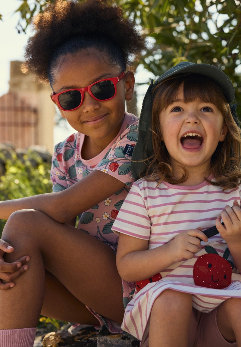 Twee kinderen zitten buiten. Het ene kind draagt een roze shirt met aardbeienpatroon en een zonnebril in het rood, en het andere kind heeft een gestreepte roze jurk met een aardbei-applicatie.