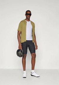 Jasnooliwkowa koszulka rowerowa z krótkim rękawem, biała podkoszulka, czarne spodenki rowerowe i białe sneakersy. Model trzyma czarny kask rowerowy.