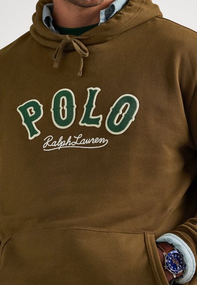 Άνδρας που φοράει καφέ φούτερ POLO Ralph Lauren με κορδόνια, από πάνω από ανοιχτό μπλε πουκάμισο, και μπλε καντράν ρολογιού στον αριστερό καρπό.