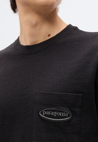 Άτομο που φοράει μαύρο t-shirt της Patagonia με μικρή τσέπη στο στήθος και κεντημένο λογότυπο, καδραρισμένο από τον λαιμό μέχρι το μέσο του κορμού.