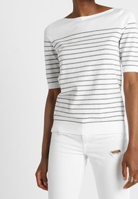 Bílý top s krátkými rukávy se šedými vodorovnými pruhy, výstřihem na loď a volným střihem. Kombinován s bílými džíny s distressed efektem.