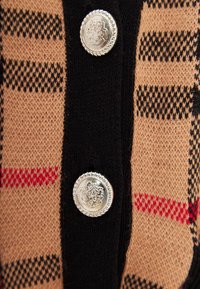 Cárdigan con un patrón de rayas beige, negro y rojo, que presenta un tejido texturizado y un botón plateado con diseño en relieve en la parte delantera.