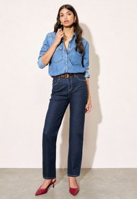 Plavi traper košulja s prednjim džepovima, tamnoplave ravne traperice i crvene cipele na visoku petu, postavljeno na neutralnoj pozadini.