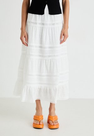 Jupe blanche brodée à volants avec un ourlet en forme de vagues, associée à des sandales plateforme orange chunky avec deux brides horizontales.