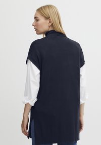 Top nero senza maniche con colletto alto, indossato sopra una camicia bianca a maniche lunghe. Presenta aperture laterali e una texture liscia.