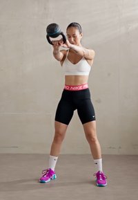 Fekete sportos rövidnadrág, fehér sportmelltartó és színes edzőcipő. Kettlebellt tartva, összpontosítva, világos beton háttér előtt.