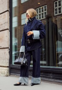 Woman wearing oversized dark denim jacket and pants with light cuffs, black pointed heels, sunglasses, and carrying a black handbag with snake-pattern handles.