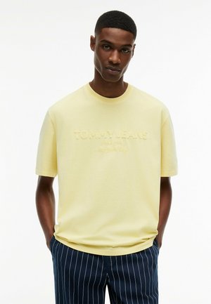 Ung mand iført en lysegul Tommy Jeans t-shirt med præget logo og marineblå bukser med nålestriber, hænderne i lommerne, hvidt ensfarvet baggrund.