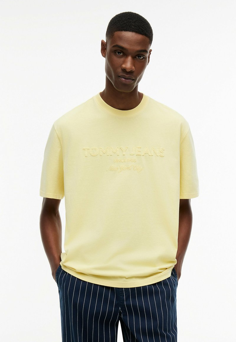 Jeune homme portant un t-shirt Tommy Jeans jaune pâle avec logo embossé et pantalon bleu marine à fines rayures, mains dans les poches, fond blanc uni.