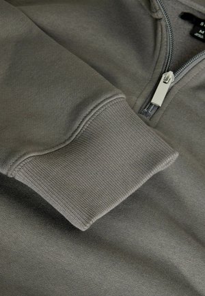 Grauer Kapuzenpullover mit Reißverschluss, der über einen gerippten Saum und eine glatte Textur verfügt. Der Reißverschluss ist aus Metall und befindet sich am Kragen.
