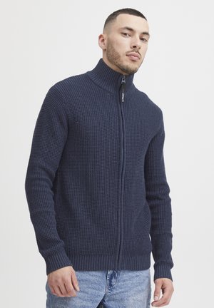 Marineblå zip-up genser med høy krage, strukturert strikkemønster, ribbestrikkede mansjetter og nederkant, samt kontrasterende svart glidelåsdetalj.