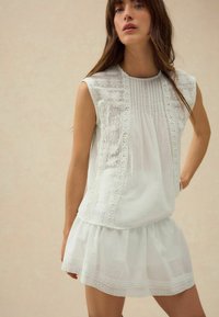 Robe blanche sans manches avec des motifs floraux brodés le long du corsage et une jupe superposée. Tissu léger et aérien avec une coupe détendue.