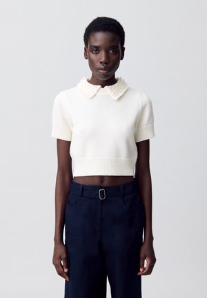Pull blanc court avec des manches courtes et un col texturé, associé à un pantalon taille haute bleu marine, muni d'une ceinture et de poches.