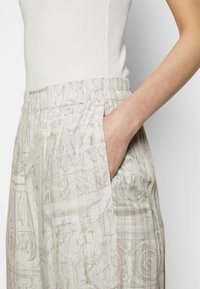 Personne portant un haut blanc côtelé et un pantalon clair à motifs avec une taille élastique, la main droite placée dans la poche du pantalon.