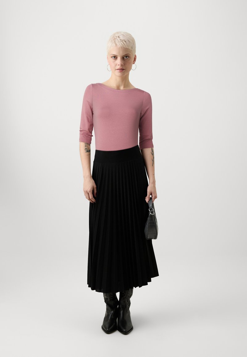 Rosa taillierte Bluse mit 3/4 Ärmeln, kombiniert mit einem schwarzen, gefalteten Midi-Rock. Die Accessoires bestehen aus einer kleinen schwarzen Handtasche und schwarzen Stiefeln.