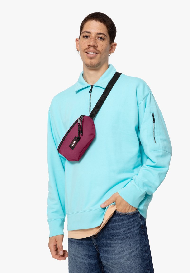 Felpa turchese con colletto a mezzo zip, indossata sopra una camicia chiara. Una borsa a tracolla bordeaux è portata, con chiusura a zip e etichetta del marchio.