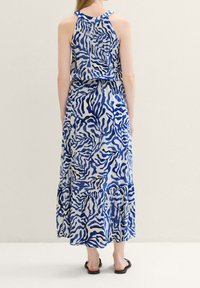 Frau in einem ärmellosen Maxikleid in Blau und Weiß mit abstrakten Wellenmustern, barfuß vor einem schlichten Hintergrund stehend.