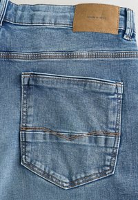 Dżinsy z denimu o jasnoniebieskim odcieniu, kontrastującym przeszyciem i naszywką z napisem "DENIM SUPPLY" na pasku. Widoczny detal tylnej kieszeni.