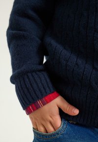 Granatowy sweter w splot kablowy z prążkowanymi mankietami, z widoczną czerwoną kratką pod spodem. Ręcznie umieszczony w kieszeni dżinsów.