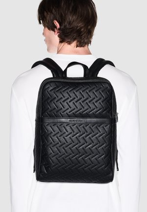 Persona che indossa una camicia bianca con uno zaino in pelle trapuntata nero con motivo geometrico e manico superiore, vista da dietro.