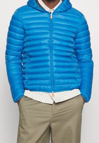 Giacca a vento blu con design a chevron orizzontale, zip frontale e cappuccio. Presenta una texture morbida e una zip a contrasto.
