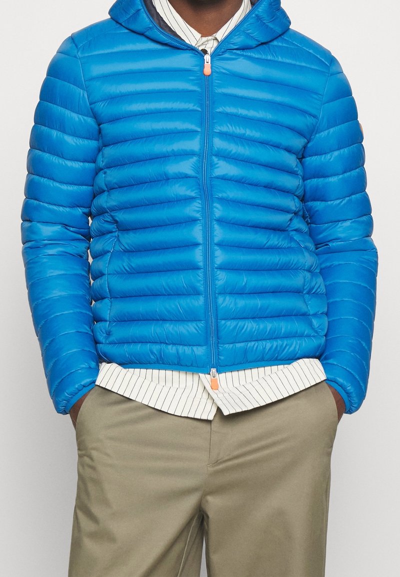 Giacca a vento blu con design a chevron orizzontale, zip frontale e cappuccio. Presenta una texture morbida e una zip a contrasto.