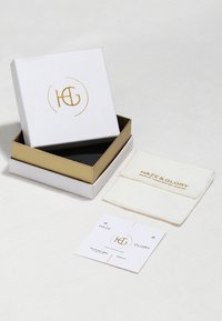 Witte doos met gouden accenten, met een "HG" logo. Inclusief crèmekleurige sieradenpouch en informatiekaart op een lichte ondergrond.
