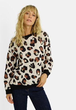 Pull en imprimé léopard beige, avec col et poignets côtelés noirs, parsemé de taches marron et crème. Texture douce, coupe décontractée.