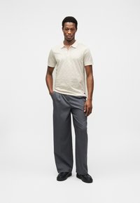 Μπλούζα πόλο με κοντά μανίκια σε ανοιχτό μπεζ χρώμα με διακριτικό μοτίβο, συνδυασμένη με γκρι παντελόνι και μαύρα παπούτσια, στέκεται μπροστά σε απλό φόντο.