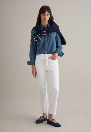 Frau trägt ein blaues Jeanshemd, weiße High-Waist-Jeans, einen marineblauen Pullover lässig über den Schultern drapiert und marineblaue flache Schuhe, steht vor einem einfarbigen Hintergrund.