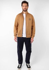 Sweat-shirt zippé marron avec col montant et motif brodé noir, associé à un pantalon marine et des baskets noires avec des accents blancs.