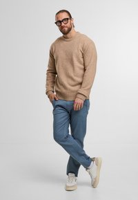 Maglione lavorato a maglia beige con scollatura rotonda e polsini a coste, abbinato a pantaloni blu chiaro. Sneakers bianche e beige completano il look.