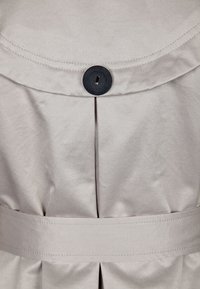 Vestido de tela beige con un botón negro redondo en el collar y un cinturón en la cintura, con un diseño plisado en la parte trasera para un detalle adicional.