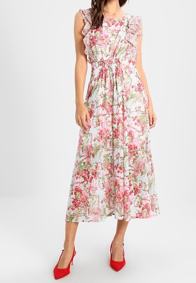 Femme portant une robe midi blanche sans manches avec un imprimé floral rose et des détails à volants, assortie à des chaussures à talons hauts rouge vif.