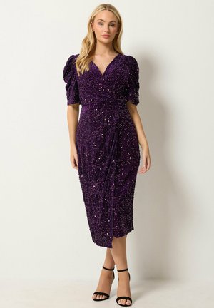 Mujer con vestido midi de lentejuelas morado con mangas abullonadas y sandalias negras de tacón alto, de pie frente a una pared lisa y clara.