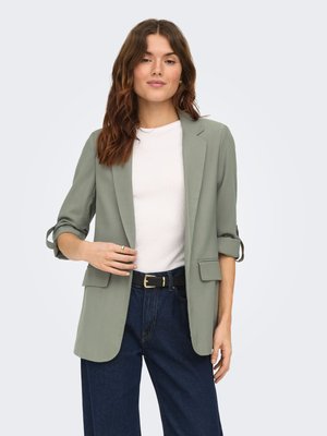 Donna con capelli castano mossi che indossa un blazer verde chiaro con maniche arrotolate, top bianco, jeans blu e cintura nera su uno sfondo semplice.