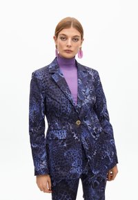 Lila Tierdruck-Anzugjacke mit tailliertem Design, langen Ärmeln und einem einzigen Knopfverschluss. Über einem lila Rollkragenpullover getragen.