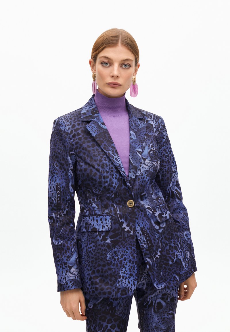 Lila Tierdruck-Anzugjacke mit tailliertem Design, langen Ärmeln und einem einzigen Knopfverschluss. Über einem lila Rollkragenpullover getragen.