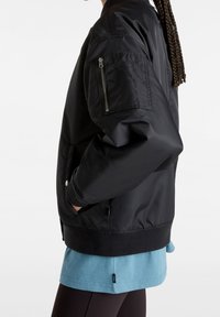 Veste bomber noire avec col et poignets côtelés. Dotée d'une poche zippée sur le bras supérieur et de poches latérales. Le matériau semble lisse et brillant.