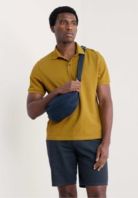 Sennepsgul polo shirt, navy sling taske og mørke shorts. Poloen har en klassisk krave, og tasken har en buet form med en strop.