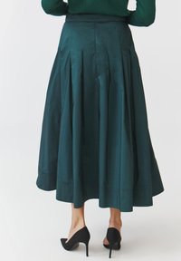 Donkergroene plooienrok gemaakt van gladde stof, met een lengte tot halverwege de kuit en een aansluitende tailleband, gecombineerd met zwarte high heels.