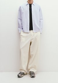 Chemise à rayures bleu clair et blanc, portée par-dessus un t-shirt noir. Pantalon ample crème et baskets gris-noir à motifs.