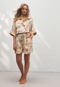 Mujer de pie descalza con pantalones cortos y camisa sueltos con un estampado tropical y náutico en tonos pastel frente a una pared blanca texturizada.