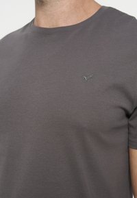 Homme portant un t-shirt gris foncé uni avec un petit logo brodé sur le côté gauche de la poitrine, visible du cou jusqu'au milieu du torse.