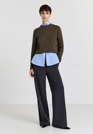 Une personne jeune portant un pull en mailles torsadées marron par-dessus une chemise boutonnée bleu clair, associée à un pantalon noir à jambes larges et des chaussures noires à bouts pointus.