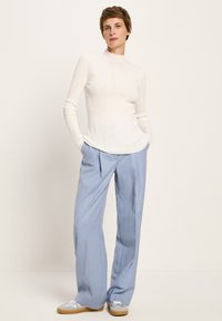 Frau steht mit Händen in den Taschen, trägt einen weißen gerippten Pullover, hellblaue weite Hosen und silberne Sneakers mit Gummi-Sohlen.